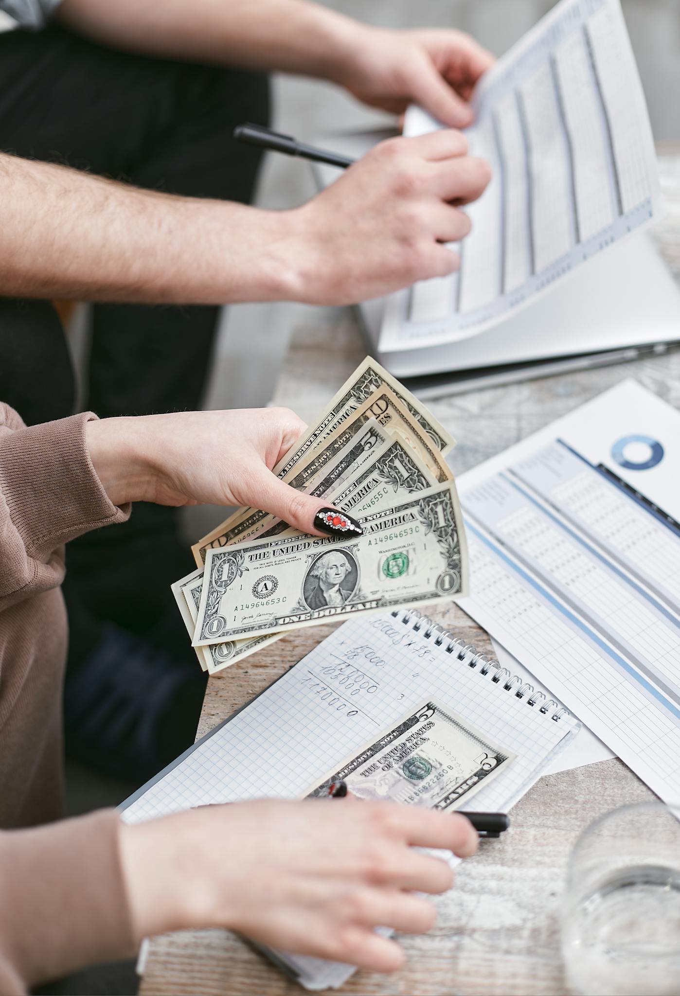 Two individuals counting dollar bills and reviewing financial documents indoors.
